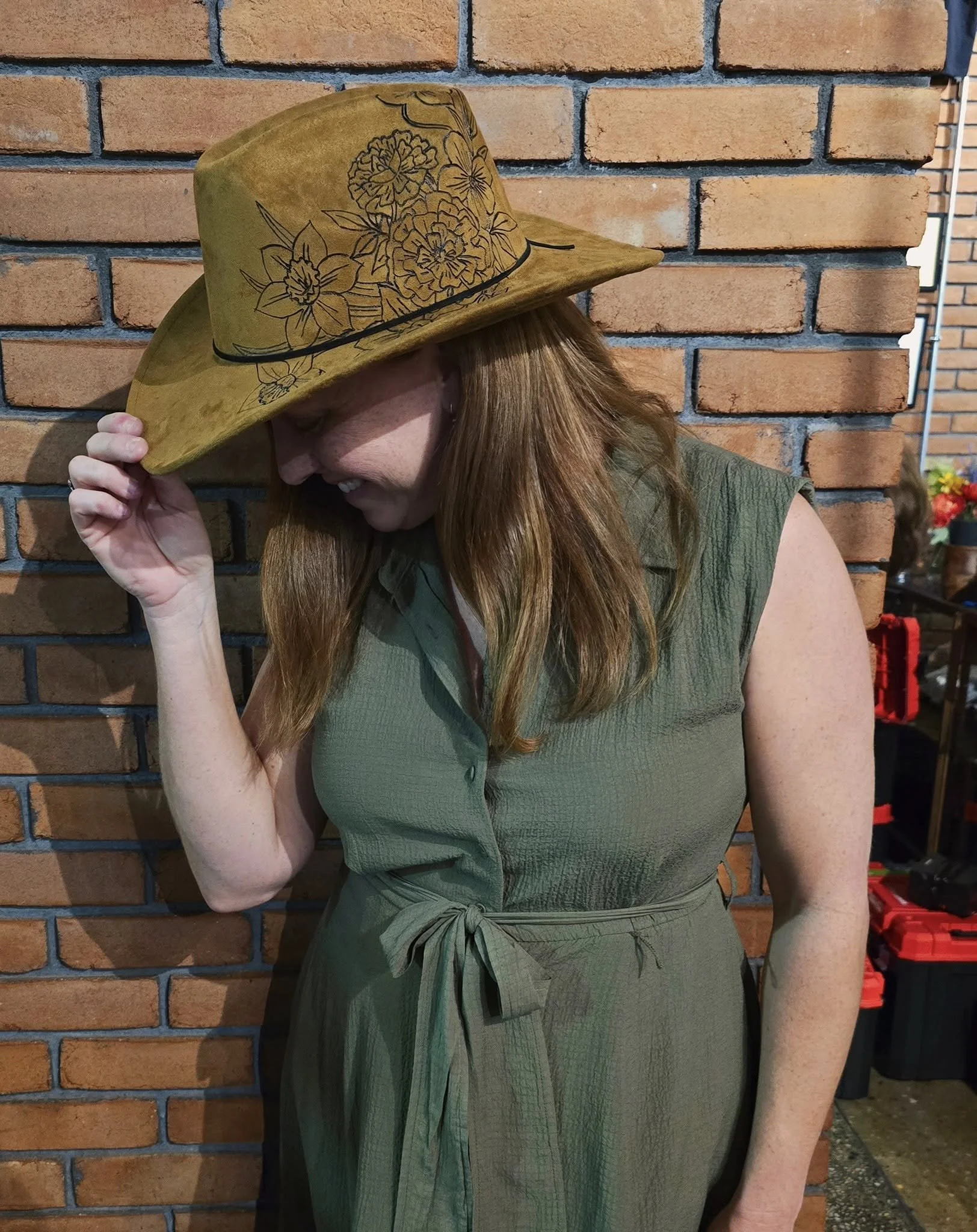 A woman with red hair is wearing a green dress and a yellow hat with black floral designs. She is holding the brim of the hat with her left hand and smiling while standing in front of a brick wall.