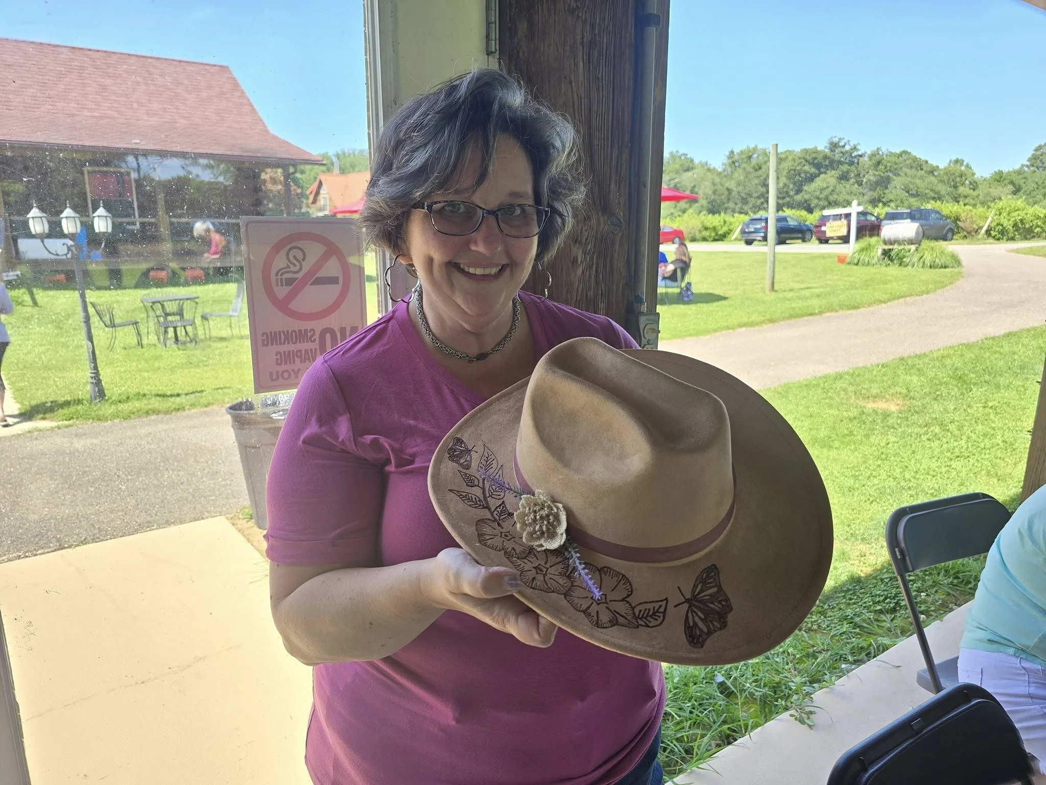 A woman with short gray hair, glasses, and a pink shirt is holding a wide-brimmed cowboy hat. She is smiling inside a building with a window looking out onto a grassy area with parked cars, trees, and outdoor seating on a sunny day.