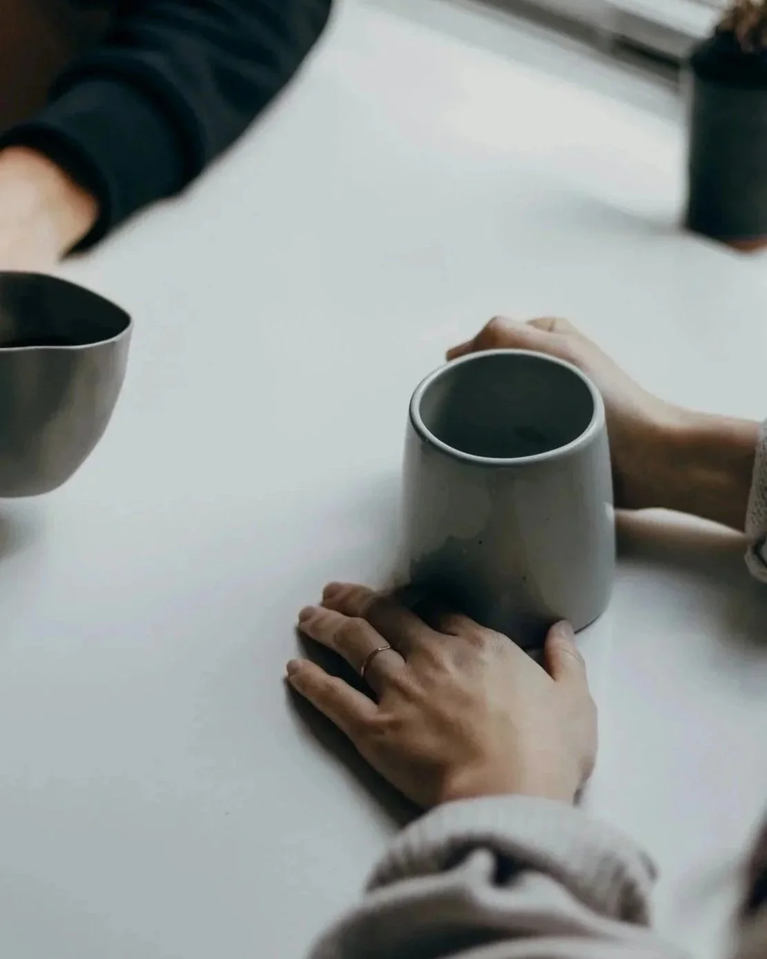 Two people holding coffee mugs at a white table, with one person's hand visible wearing a ring.