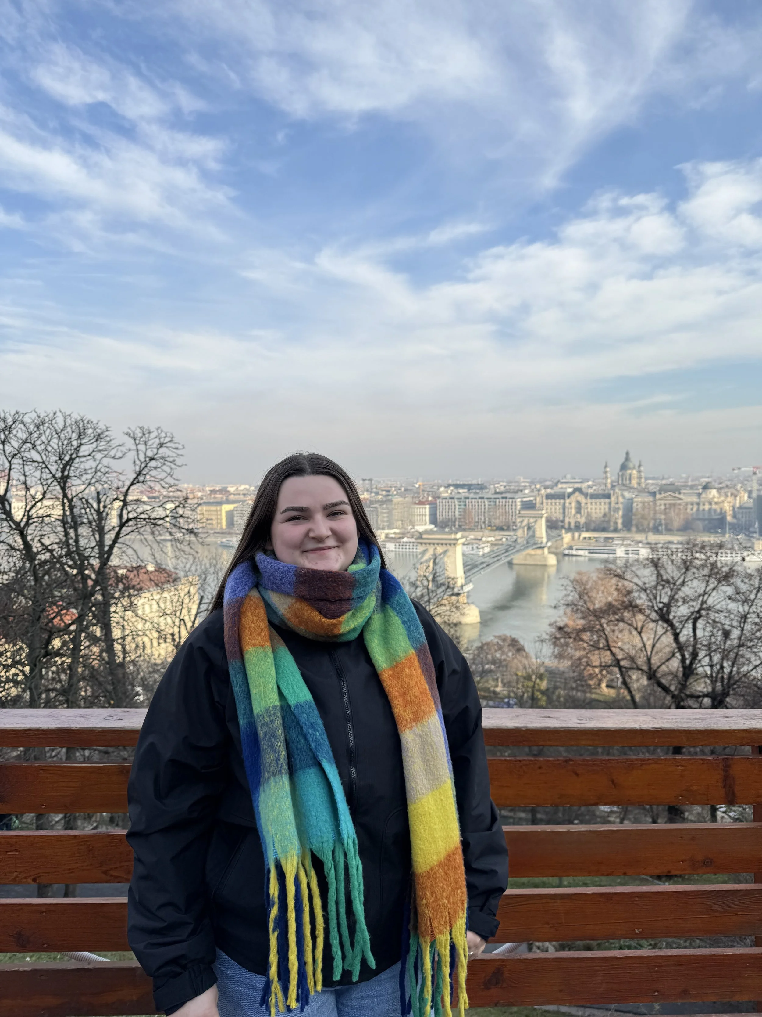 Becky a white woman in stood infront of a low fence overlooking the London skyline. She's wearing a blue and orange checered scarf and a black puffer. Her hair is long, black and straight.