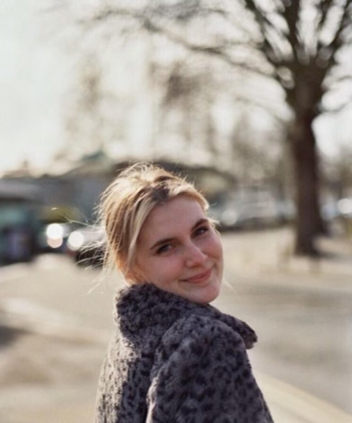 Isobel a white woman is stood body turned away from the camera but head turned to face us- she's wearing furry leopard print coat with blonde hair tied back. In the background a blurry view of a curved street with a leafless tree.