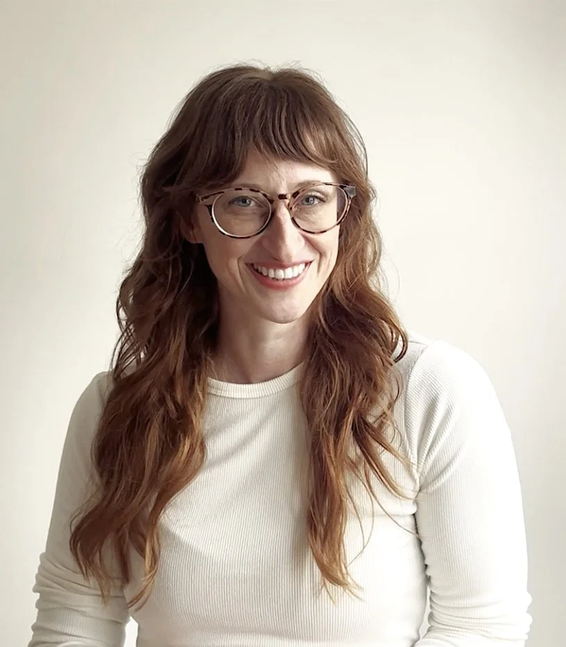 A woman with red, wavy hair and glasses smiling at the camera, wearing a white long-sleeve shirt, against a plain light-colored background.