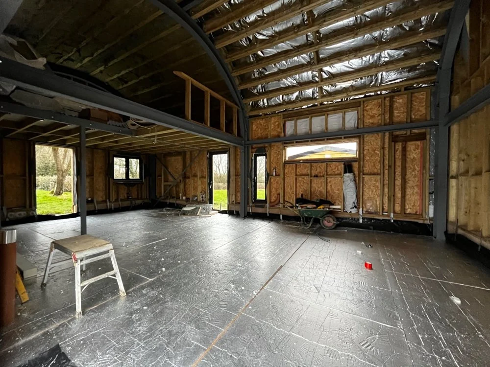 Interior of a house under construction with exposed wooden framing and metal beams. The floor is unfinished, and there are windows and a door opening showing the outside. Construction tools and materials are visible inside.