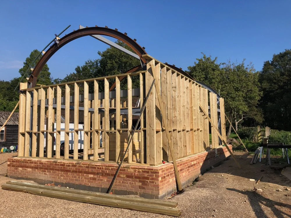 Construction of a building with a brick foundation and wooden frame, with an arched wooden structure being installed on top, surrounded by trees and clear blue sky.