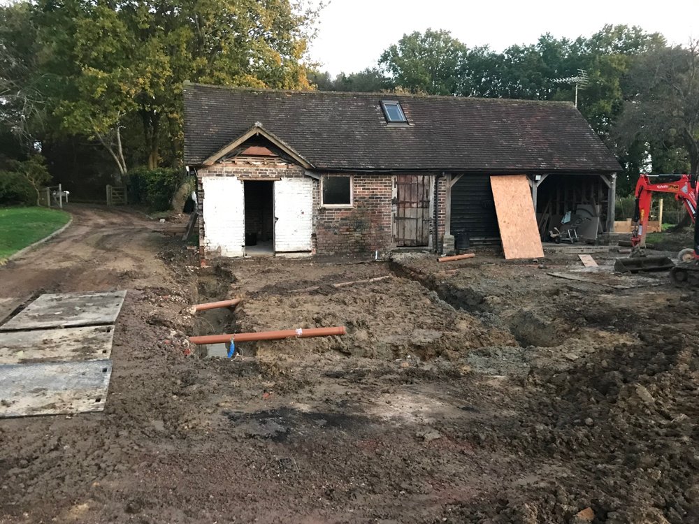 A house undergoing construction or renovation, with visible trenching and piping on the ground in front of the house, and construction equipment nearby.