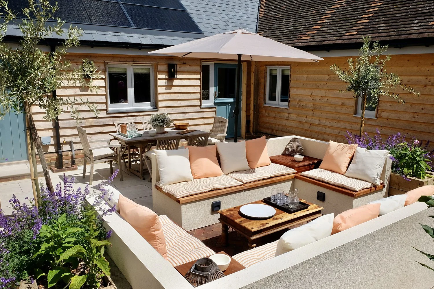 Decorative outdoor patio with white sectional sofas, cushions, and throw pillows, a wooden coffee table with glasses and a tray, a dining table with chairs, potted plants with purple flowers, a large patio umbrella, and a wooden house with windows an