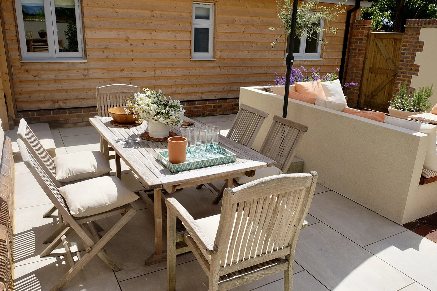 Outdoor patio with wooden dining table and chairs, a tray with glasses and a flower vase, a built-in bench with cushions, and a wooden house wall in the background.