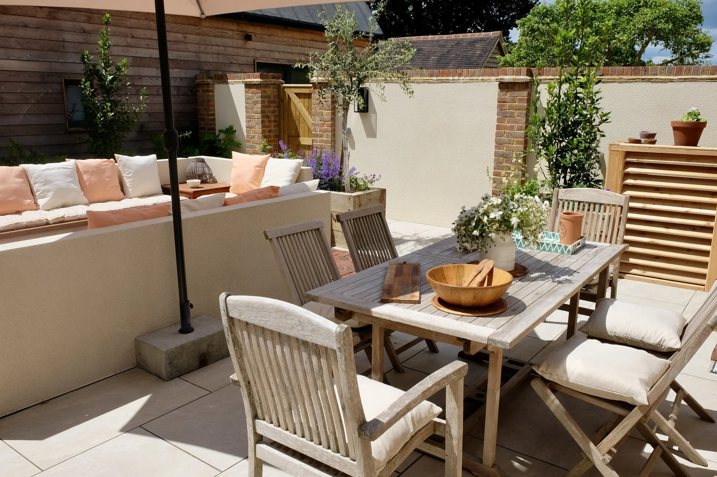 An outdoor patio area with a wooden dining table and chairs, a cushioned seating area with pillows, potted plants, and a fence with brick accents, under a sunny sky.