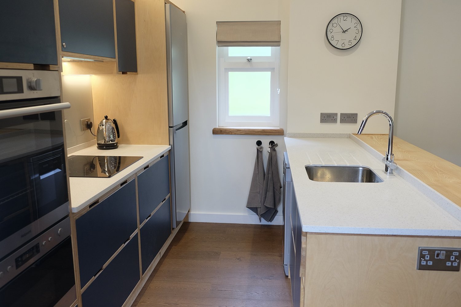 A modern kitchen with blue cabinets, a white countertop, a stainless steel sink, a kettle, an oven, a refrigerator, and a window with a beige roller blind. There is a wall clock and two electrical outlets above the sink area.