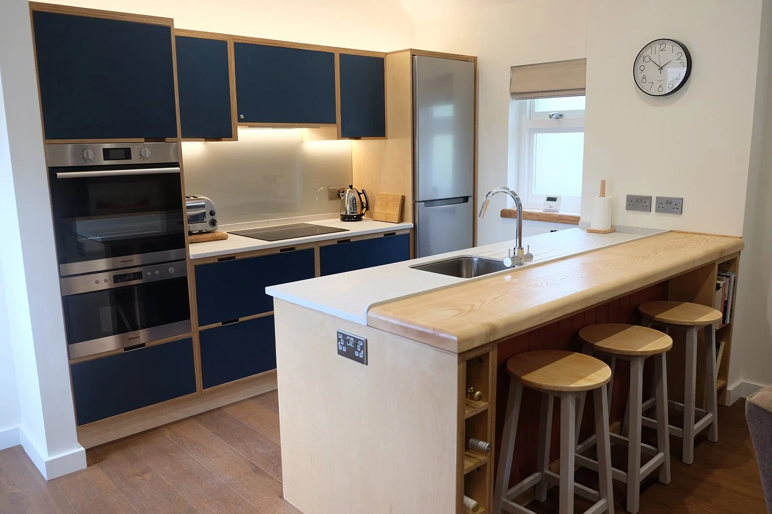 Modern kitchen with blue cabinets, stainless steel appliances, a white countertop, a wooden breakfast bar with three stools, and a clock on the wall.