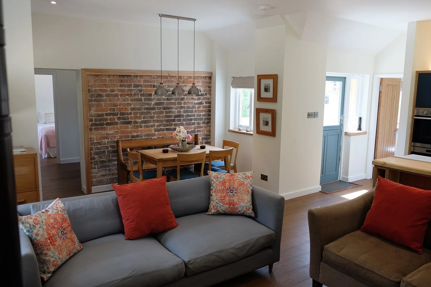 Living room with gray and brown sofas, decorative pillows, wooden dining table with flowers, brick accent wall, and a hallway leading to other rooms.