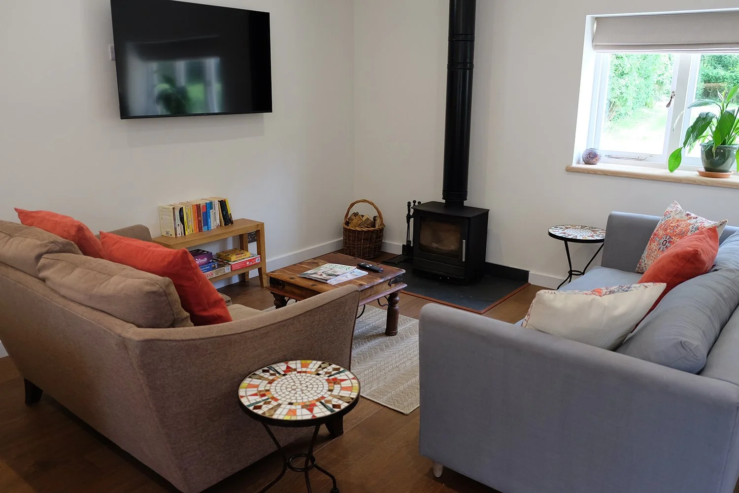 Living room with two sofas, one beige with red pillows and one grey with orange and patterned pillows, a wooden coffee table, a small colorful mosaic side table, a wall-mounted flat-screen TV, a cast iron wood stove, a window with a plant and decorat