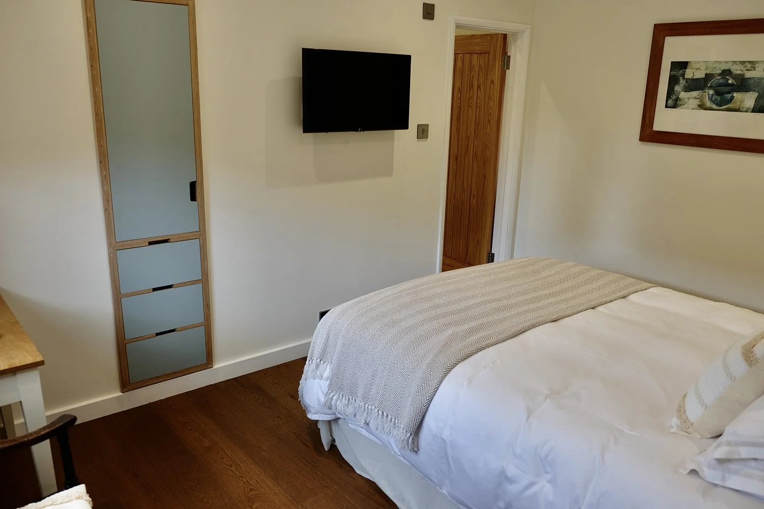A bedroom with a bed, a framed picture on the wall, a flat-screen TV mounted on the wall, a partially visible chair, and a door to a closet or another room.