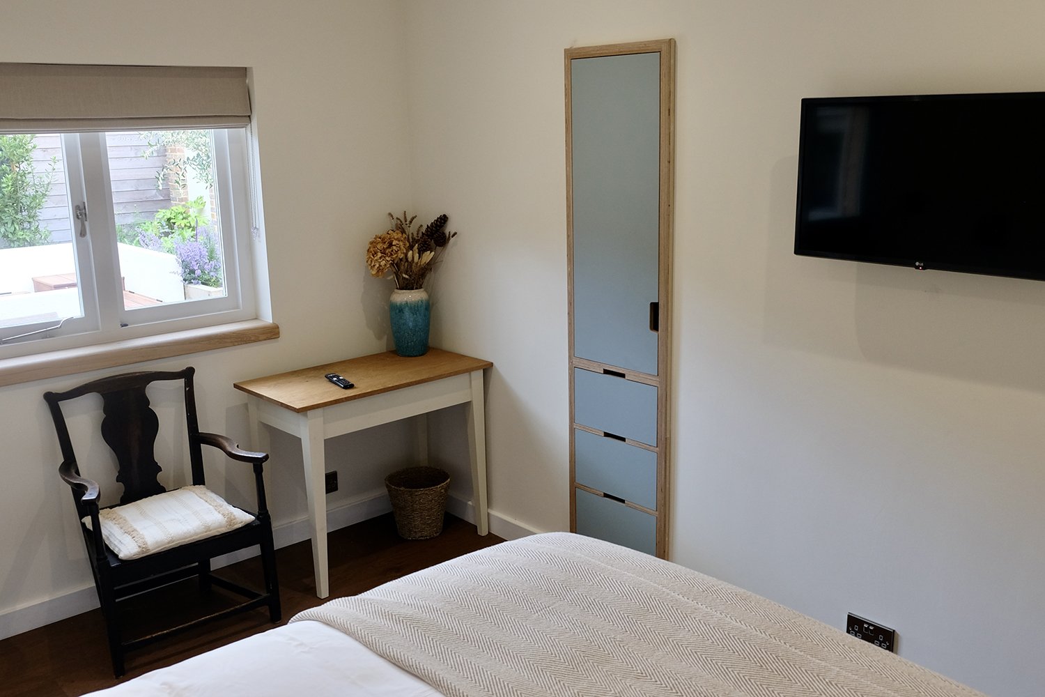 A bedroom corner with a bed, a window with a beige roller blind, a black wooden chair with a cushion, a small white desk with a phone and a vase of dried flowers, a wicker basket, a light blue wardrobe with wooden frame, and a wall-mounted TV.