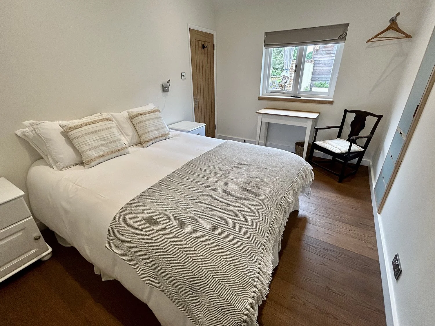 A cozy bedroom with a large bed with white and beige bedding and three matching pillows. There are two white nightstands on either side of the bed, and a small wooden desk and black wooden chair near the window. The window has a beige roller shade an