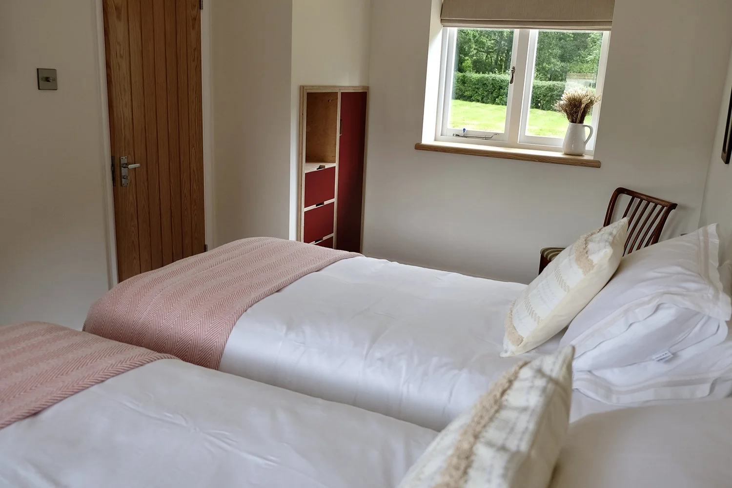 A cozy bedroom with two single beds, each with white sheets, beige pillows, and pink blankets. There is a wooden chair next to the beds, a window with a view of green grass and trees, and a small red and wood bookshelf. A white vase with dried flower