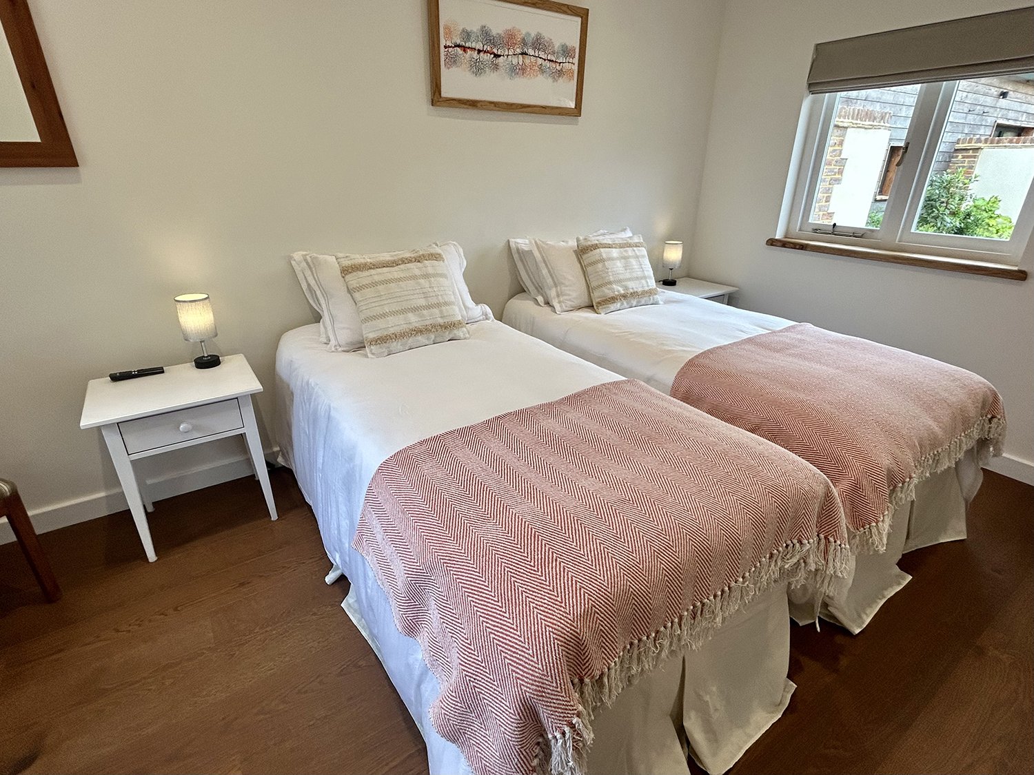 A bedroom with two single beds pushed together, each with white bedding, striped pillows, and pink blankets at the foot of the beds. There are nightstands with lamps on each side, a window with a view outside, and artwork hanging on the wall.