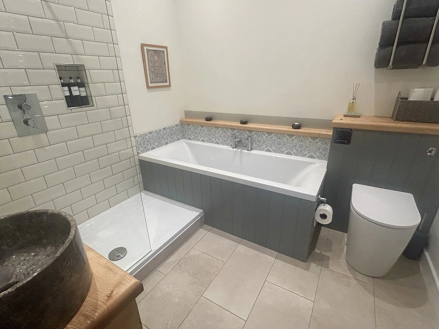 Bathroom with walk-in shower, bathtub, toilet, and wooden shelves holding decorative items, with tiled walls and beige floor tiles.