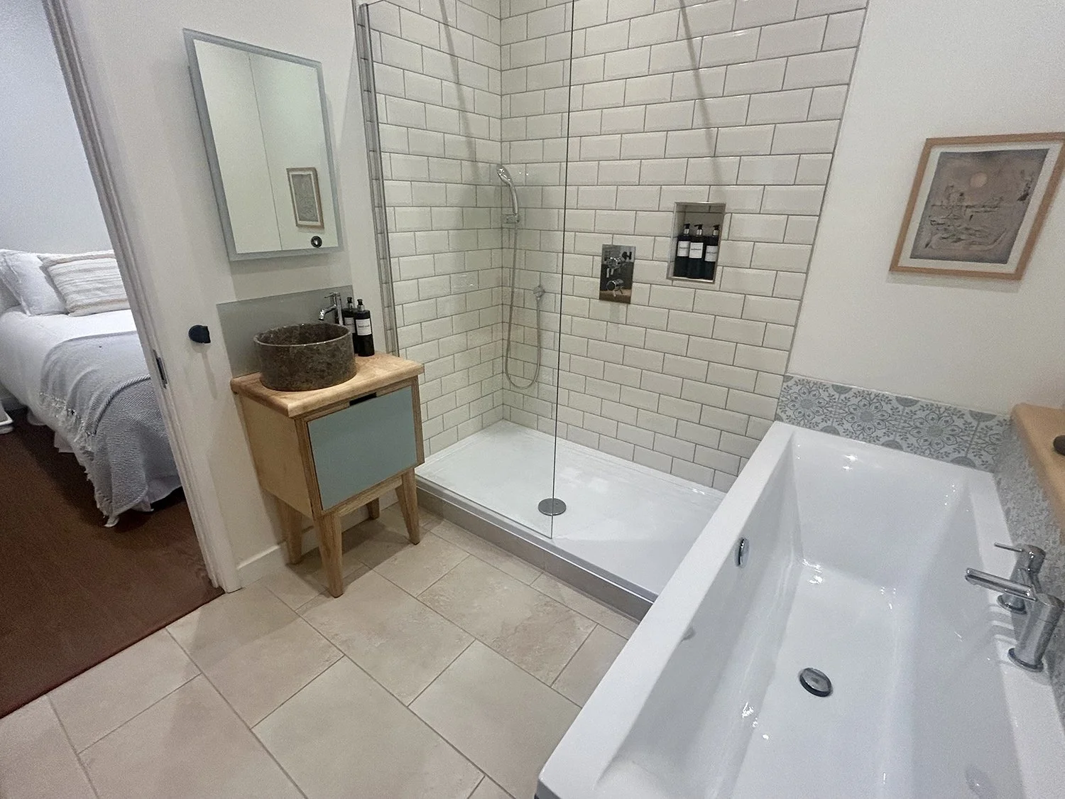 A bathroom with a walk-in shower with white brick tiles, a glass door, a hand shower, and built-in recessed shelves with toiletries. There is a white bathtub with a tiles and decorative border on the right, a wooden vanity with a stone sink and toile