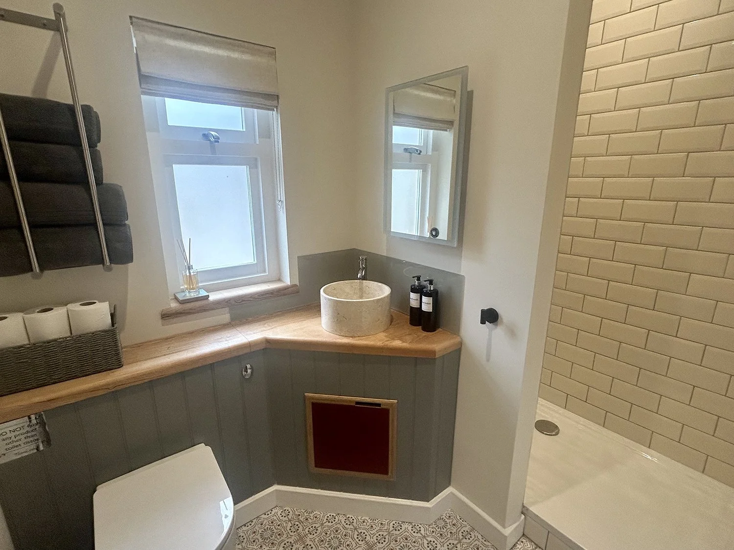 A small bathroom with a wooden countertop, a round stone sink, black soap bottles, a mirror, a window with a blind, a wall-mounted shelf with folded towels, and a shower area with beige brick tiles.