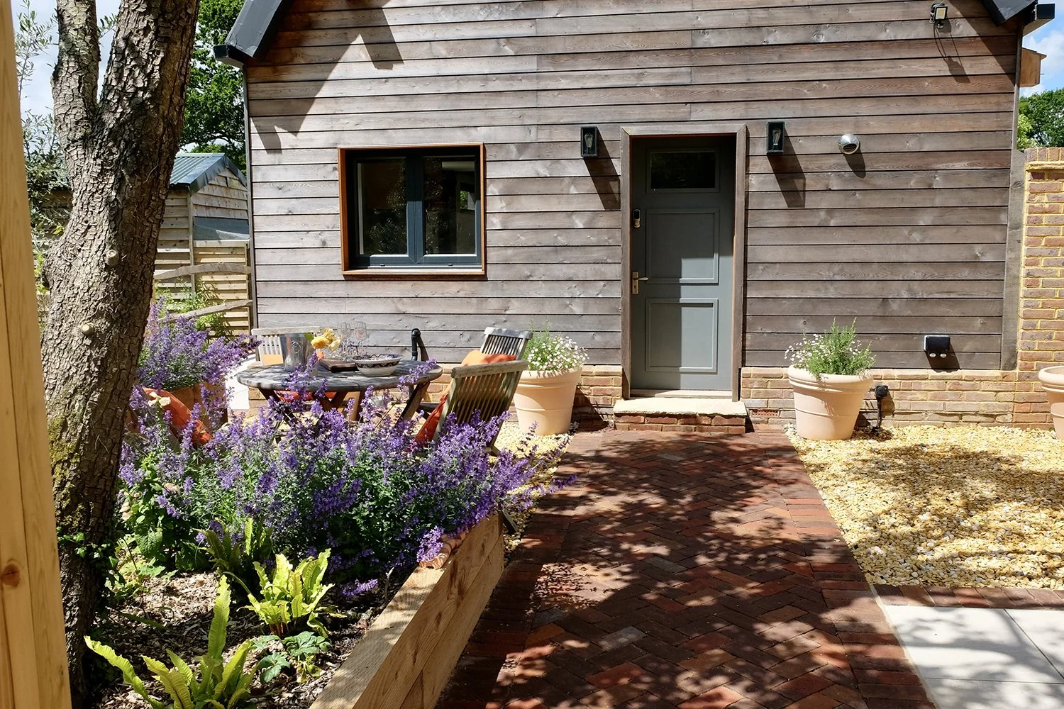 A backyard patio with a brick walkway, potted plants, a table with chairs, and a wooden house with a gray door and window.