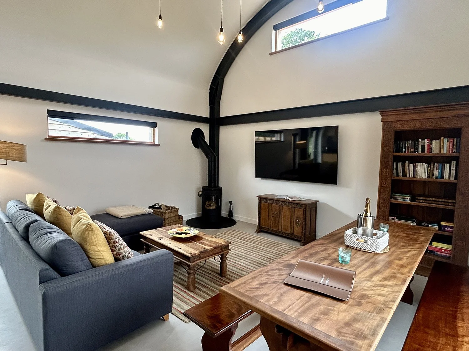 Living room with a gray sofa, yellow and patterned cushions, wooden coffee table, wood-burning stove, flat-screen TV, wooden cabinet, bookshelf, and a dining table with a laptop, glasses, and decor items. Ceiling with hanging light bulbs and narrow h