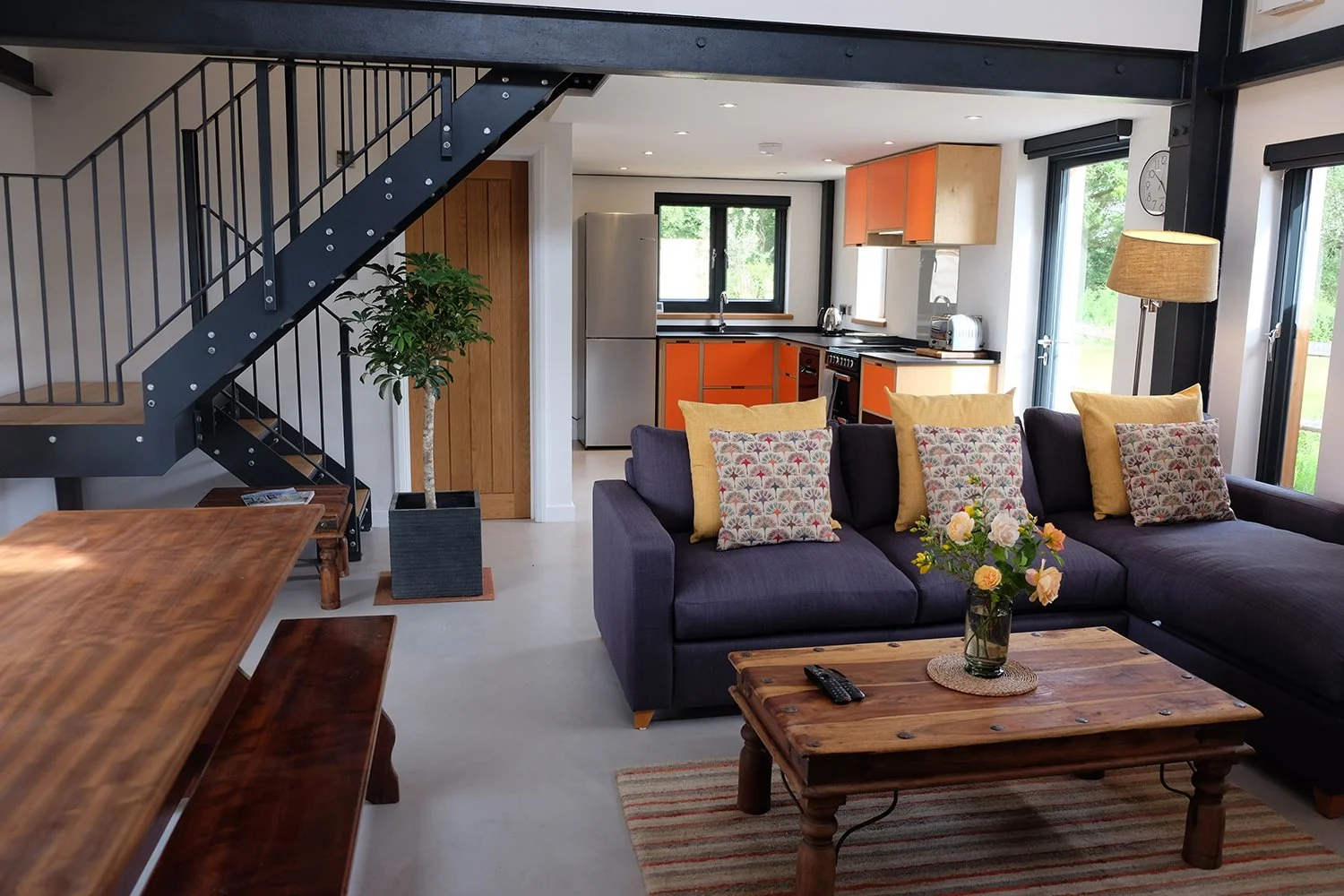 Open-plan living room and kitchen area in a modern home, featuring a dark purple sofa with decorative pillows, a wooden coffee table with a vase of flowers and remote, a large window, and a kitchen with orange cabinets, a silver refrigerator, and bla