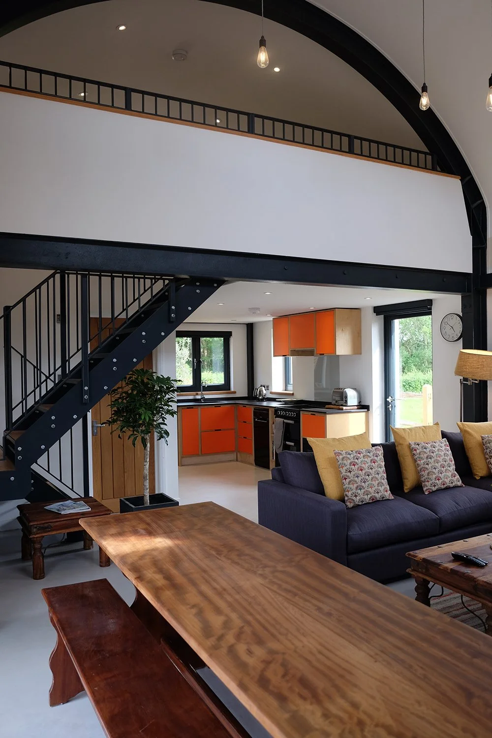 Open-concept living and kitchen area with black staircase, orange cabinetry, dark sofa with colorful pillows, wooden table, large windows, and a loft area.