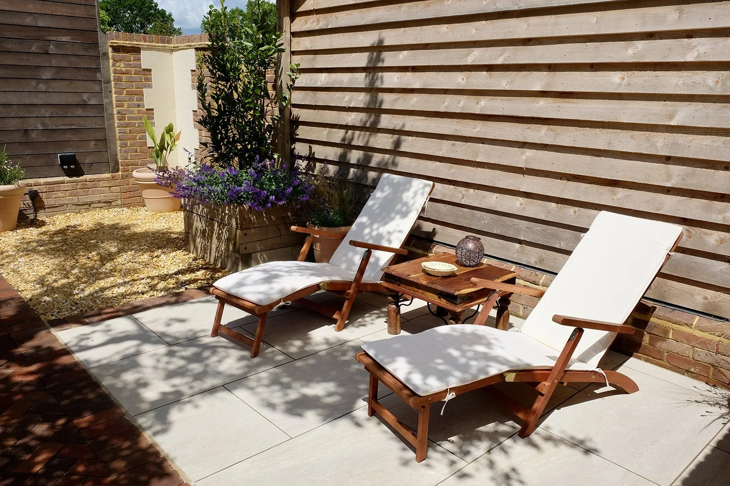 Backyard patio with two white cushioned lounge chairs, a small wooden side table, potted plants, and a wooden fence with shadow patterns.