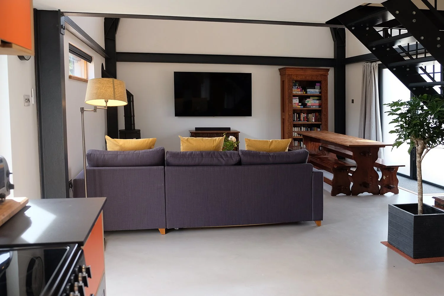 Living room with purple sofa, wooden dining table, bookshelf, mounted TV, staircase, and indoor plant in a modern home
