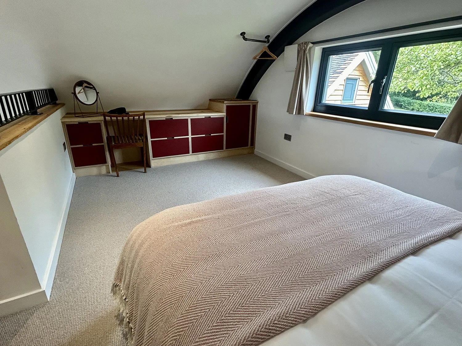A bedroom with a bed covered in a beige blanket, a window with beige curtains showing greenery outside, a built-in cabinet with red doors, a wooden chair, and a mirror on the cabinet.