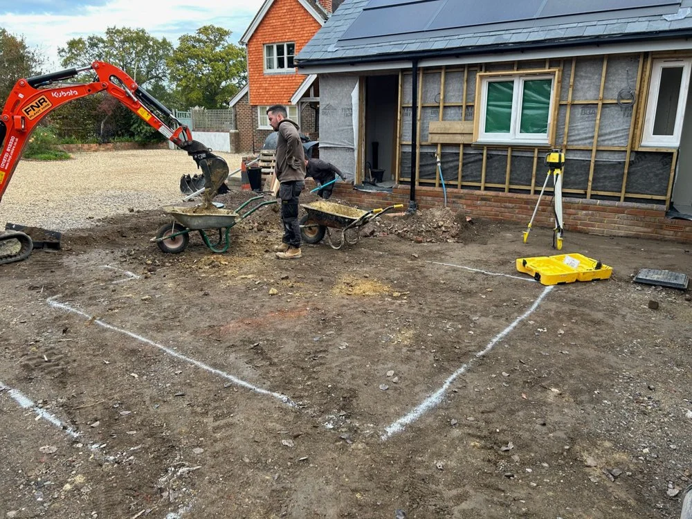Construction workers preparing the ground for a new addition or patio, with a small excavator, wheelbarrows, and surveying equipment on site.