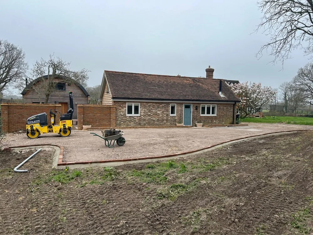 A house with a brick exterior and a tiled roof, surrounded by a partially constructed gravel driveway, construction equipment, and a wheelbarrow in the yard.