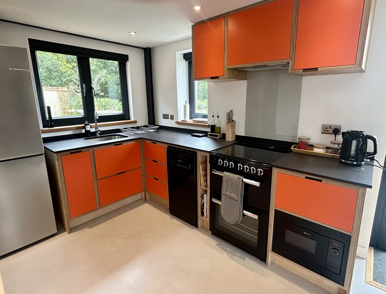 Kitchen with black countertops, orange cabinet doors, stainless steel appliances, two windows, and a view of greenery outside.