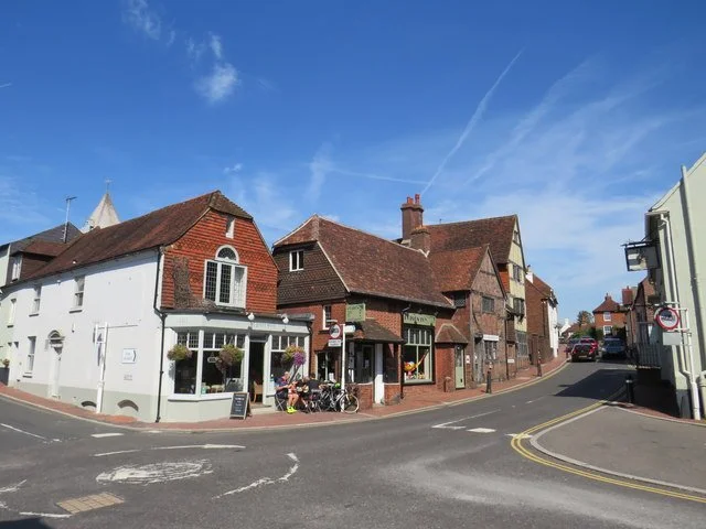 A street corner with several brick buildings, one with a white facade, cafes or shops, bicycles parked outside, under a blue sky with some clouds.