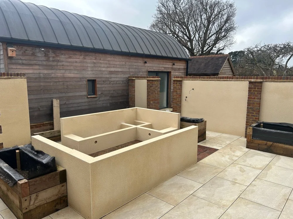 Construction site of an outdoor kitchen with a beige stucco structure, empty planters, and a wooden privacy wall, with trees and a shed in the background.