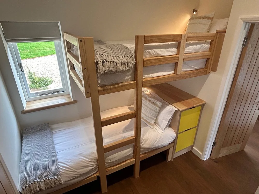 A wooden bunk bed with three sleeping areas in a room, with a window showing a grassy yard outside.