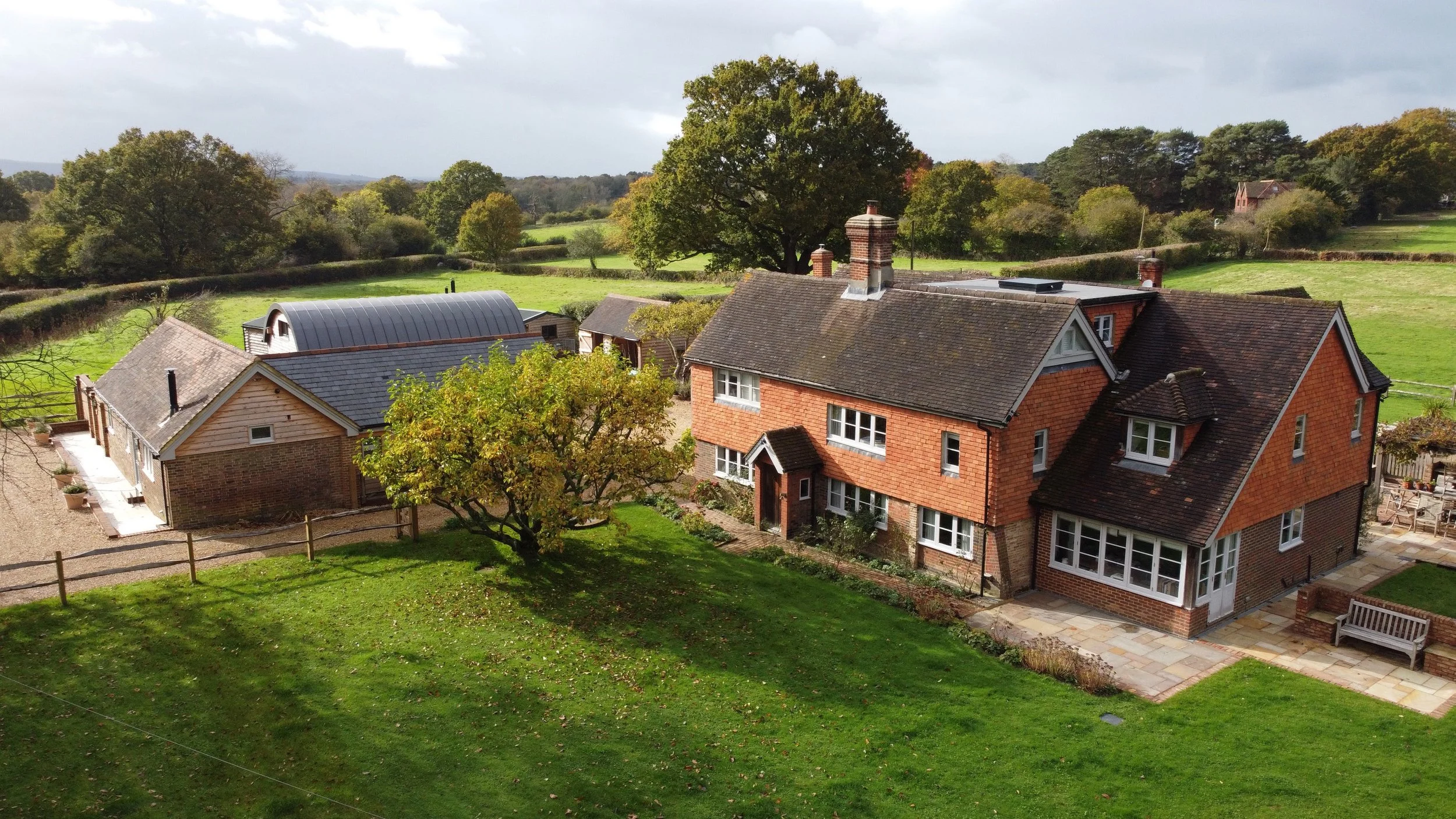 Overlooking a house with a brick exterior, tiled roof, and multiple chimneys, surrounded by green lawn and trees, with open fields and distant trees in the background.