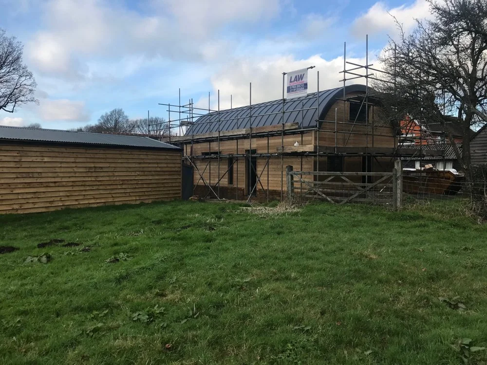 A house under construction with wooden walls, scaffolding, and a curved roof, surrounded by a green grassy yard.