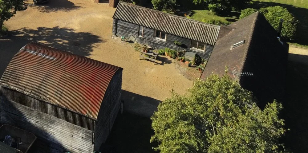 Aerial view of a rustic farmyard with two wooden buildings, one with a corrugated metal roof and the other with a tiled roof, surrounded by trees and a dirt ground, with some potted plants and garden decorations.