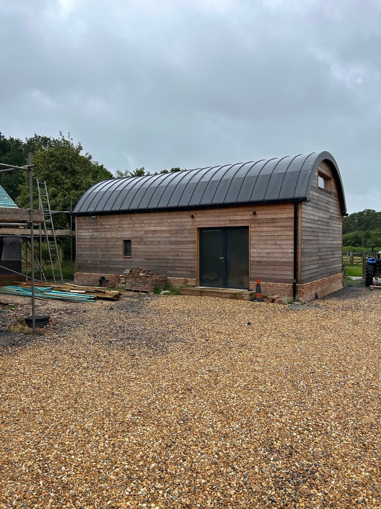 A wooden barn with a curved metal roof sits on a gravel lot, with some construction materials and scaffolding nearby.