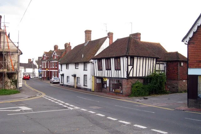 A street corner with a white house featuring black timber framing, other brick houses, a sidewalk, and a curved road.
