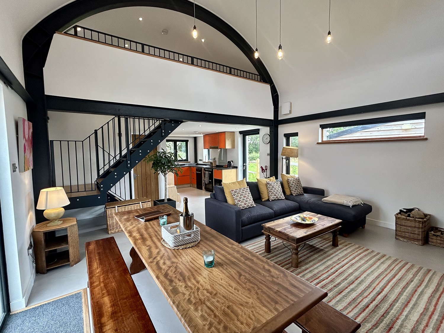 Open-concept living room with black staircase leading to an upper level, living area with dark gray sectional sofa, wooden coffee table, and striped rug, and a kitchen with orange cabinets in the background, letting in natural light from multiple win