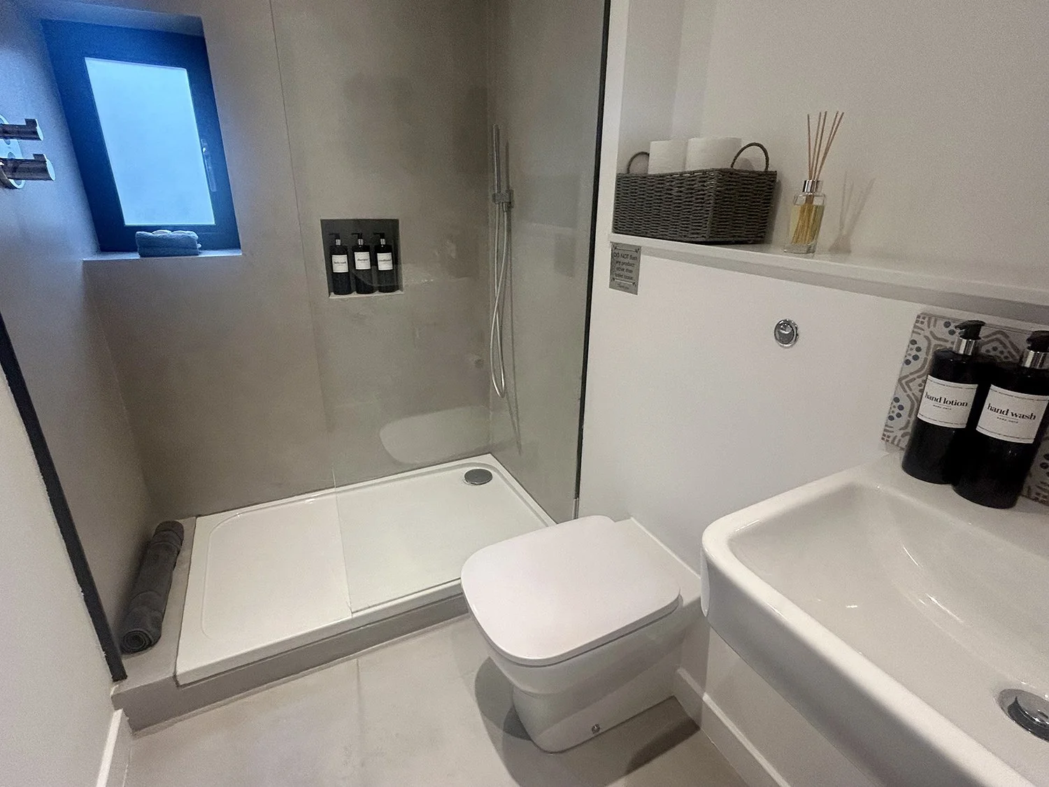 Small modern bathroom with a walk-in shower, a toilet, and a sink. The shower has stone walls, a window with a rolled towel, and bottles of toiletries. The sink has two bottles of hand soap and hand lotion. There are shelves with towels and reed diff