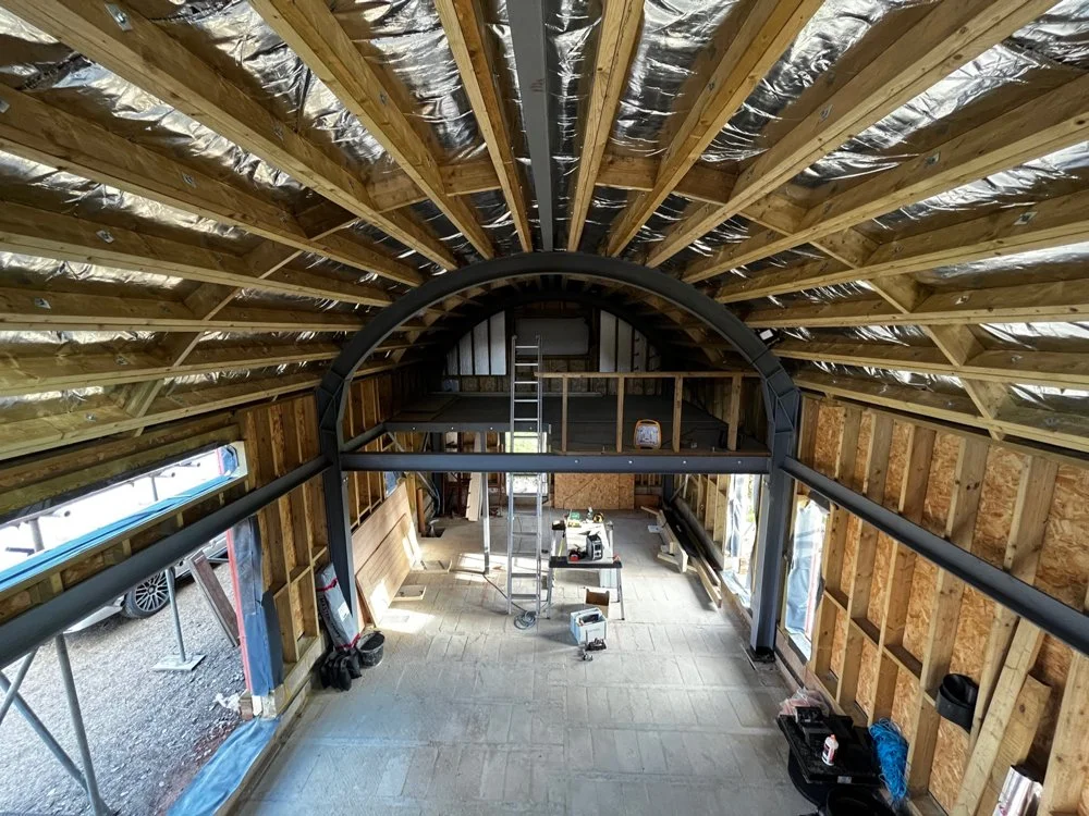 Interior of a building under construction with exposed wooden framing, metal ductwork, ladder, and construction tools visible.