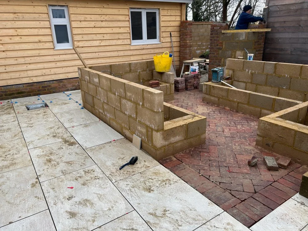 Construction of a brick patio or garden wall outside a house. Workers building brick structures, with tools and materials around.
