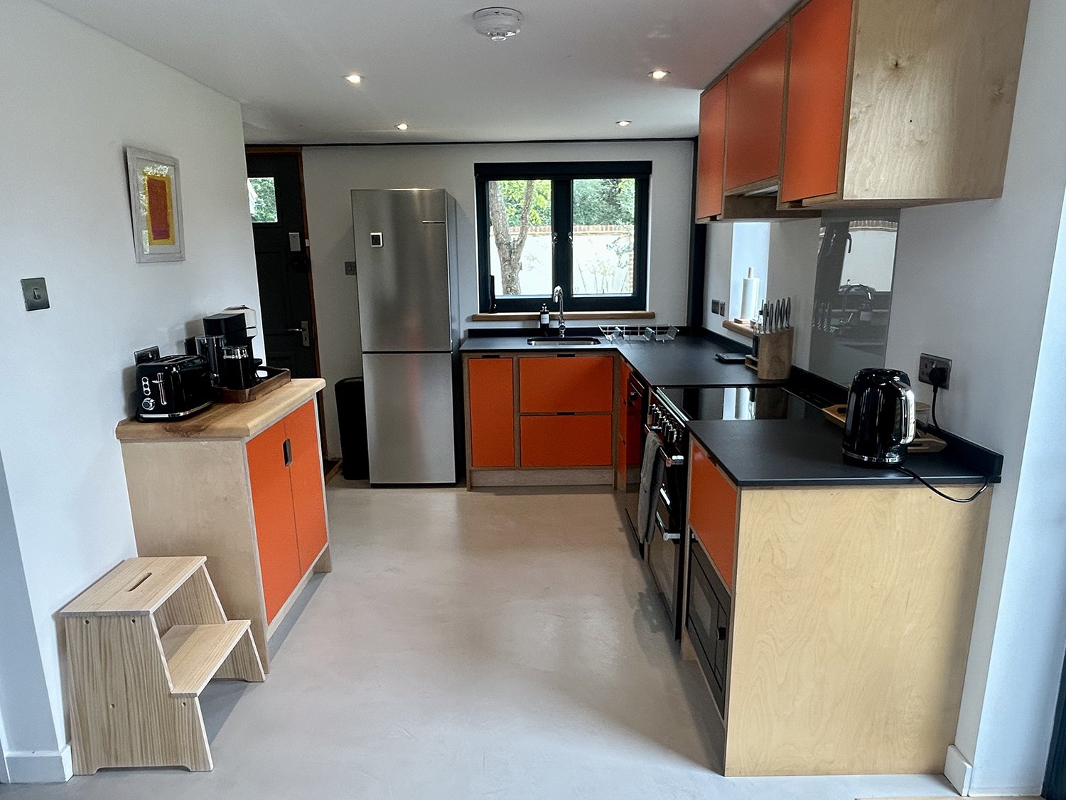 Modern kitchen with orange and natural wood cabinets, black countertops, stainless steel refrigerator, and black kitchen appliances. There is a window over the sink, and a small wooden step stool in the corner.