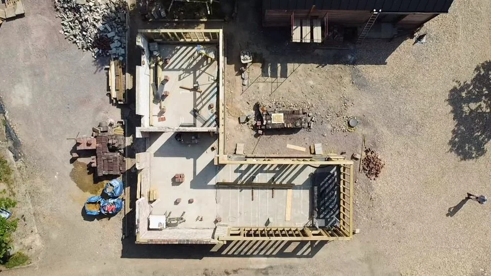 A bird's eye view of a construction site showing two partially built floors with wooden framing, construction tools, and materials, with some workers present.