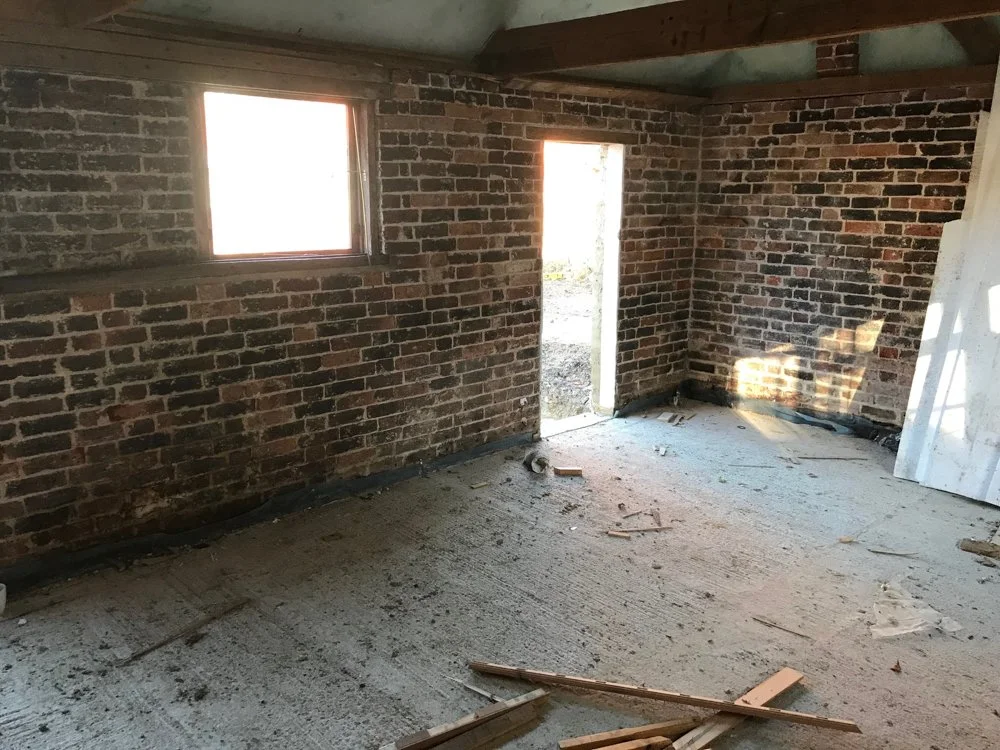 Interior of a room under construction with brick walls, an open doorway, a window, unfinished concrete floor, and construction debris.