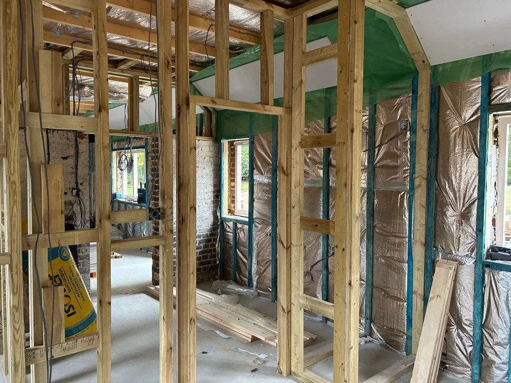 Interior view of a house under construction with exposed wooden framing, insulation, and partially completed walls.